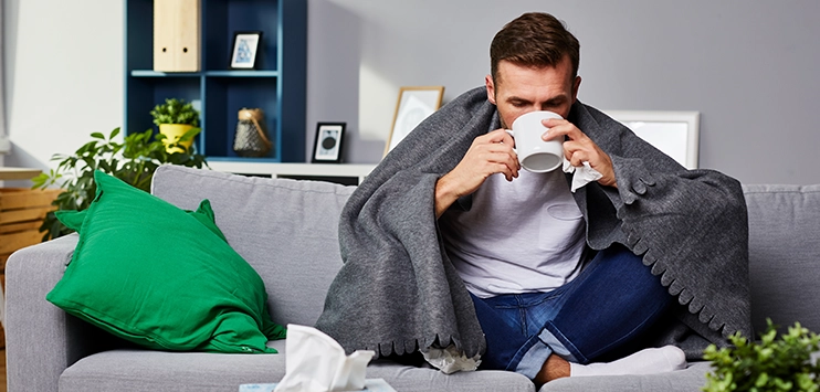 Erkälteter Mann auf dem Sofa mit Tasse und umgeben von benutzten Taschentüchern